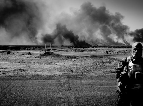 Iraqi Special Forces soldier during advance on Mosul with oil installations blown up by Daesh burning near Qayyarah. Standing in the foreground with billowing smoke in the distance over the desert.