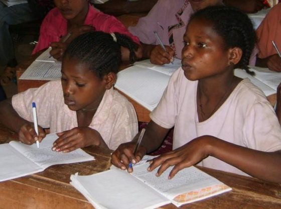 Two Ethiopian girls in class in a school supported by LCDI, Wolaita Zone, Ethiopia, 2015. Both are writing in jotters. One pays close attention to what the teacher is saying.