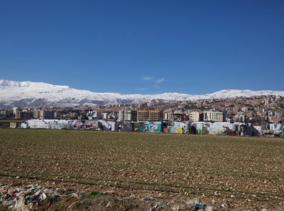 An informal tented settlement of Syrian refugees in the Bekaa Valley, Lebanon. A series of temporary shacks are in the foreground next to a ploughed, rocky field. The apartment blocks of a town are in the background. Snow covered hills are on the horizon.