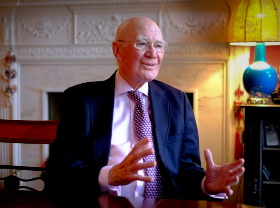 Menzies Campbell at home in Edinburgh. He faces the camera smiling. He is wearing a blue suit, shirt and tie. His arms bent and apart.
