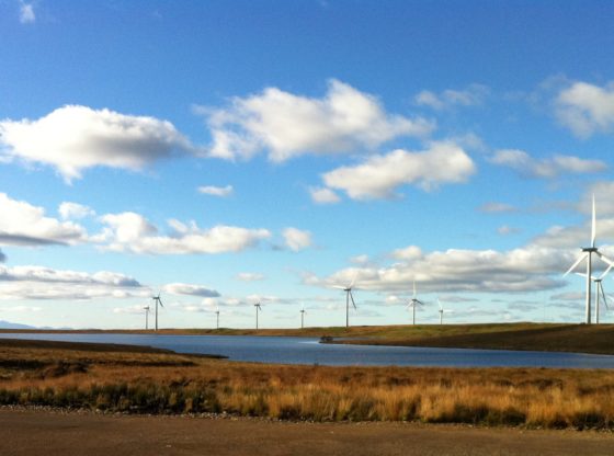 Whitelee wind farm with Arran in the background. Wind turbines dot a flat landscape beneath an open blue sky.