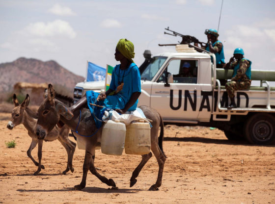 A woman rides a donkey loaded with water jerry cans, while UNAMID troops from Tanzania conduct a routine patrol in the camp for internally displaced persons (IDP) in Khor Abeche, South Darfur. In the foreground a woman rides a heavily laden donkey with a UN armed truck in the background.