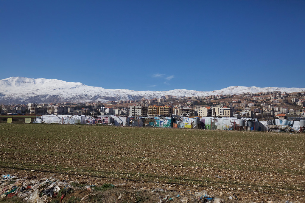 An informal tented settlement of Syrian refugees in the Bekaa Valley, Lebanon. A series of temporary shacks are in the foreground next to a ploughed, rocky field. The apartment blocks of a town are in the background. Snow covered hills are on the horizon.