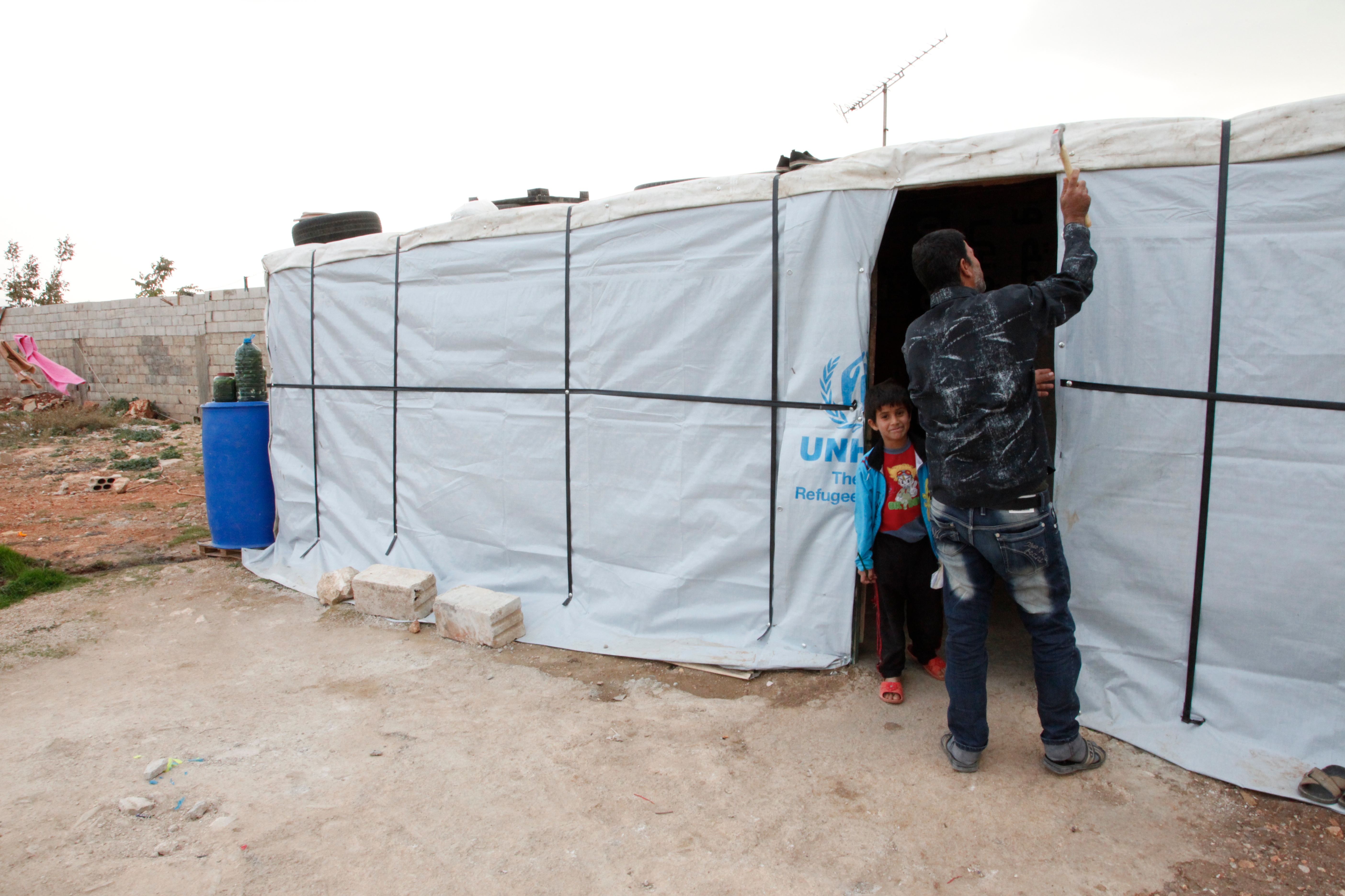 A man ties up the cloth entrance to his UNHCR tent. He has his back to the camera. His son peaks out at his side and smiles at the camera.