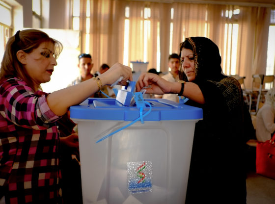 A Kurdish woman puts casts her ballot. Another Kurdish woman holds the cover of the voting slot open.