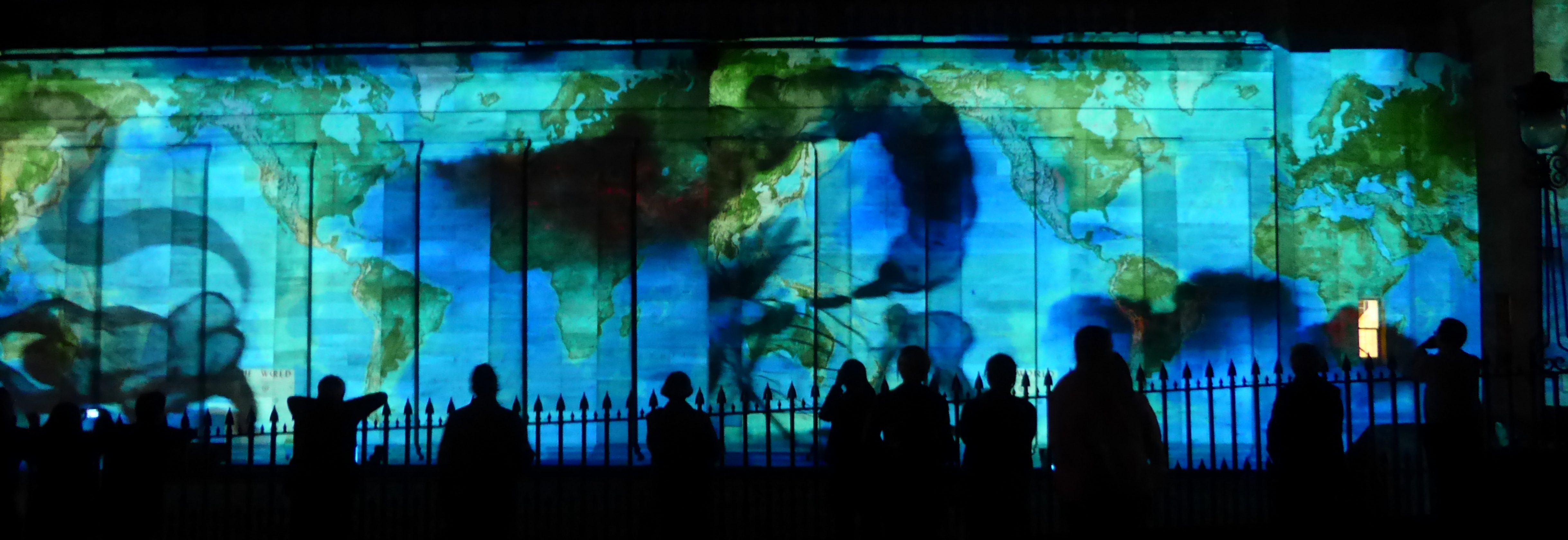 "In Search of Vanished Blood". Projection at the Edinburgh Fringe Festival. A world map overlaid with black swirls is projected onto a stone building, with people's silhouettes outlined against the light.