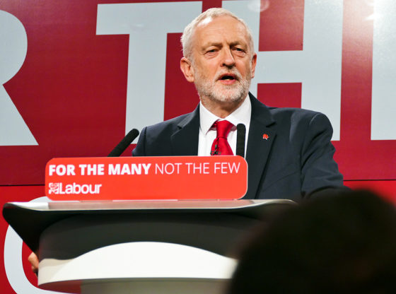 Labour leader Jeremy Corbyn stands on a stage, speaking to his audience ahead of the 2017 UK General Election. He wears a dark suit, white shirt and red tie. On the plinth he is standing at is the slogan: For The Many, Not The Few'. LABOUR'.