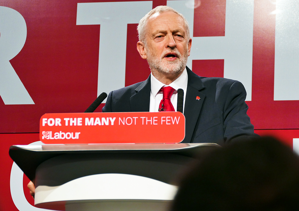 Labour leader Jeremy Corbyn stands on a stage, speaking to his audience ahead of the 2017 UK General Election. He wears a dark suit, white shirt and red tie. On the plinth he is standing at is the slogan: For The Many, Not The Few'. LABOUR'.