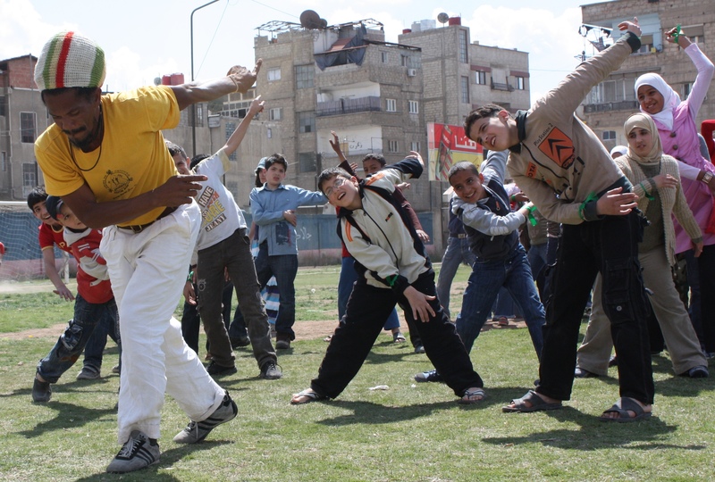 A capoeira class for children in Damascus affected by the on-going Syrian conflict. A man leads a group of children in a dance on a football pitch, with apartment buildings in the background.