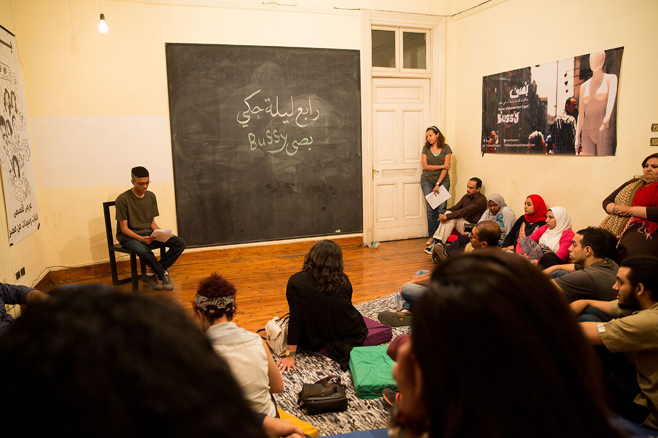 A storytelling night at Bussy in Damascus, which creates safe spaces for Egyptians to recount their experiences. In a bright room with Arabic written on a large blackboard, a young man reads aloud to a group of listeners seated on the floor.